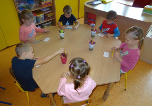 Sześcioro dzieci siedzi przy stoliku na którym rozłożone są kubeczki z kolorowymi kredkami. Przed każdym dzieckiem leży kartka z obrazkami kredek ułożonymi w różne kompozycje. Dzieci sięgają do kubeczków po kredki i próbują odtworzyć układ kredek z obrazka.